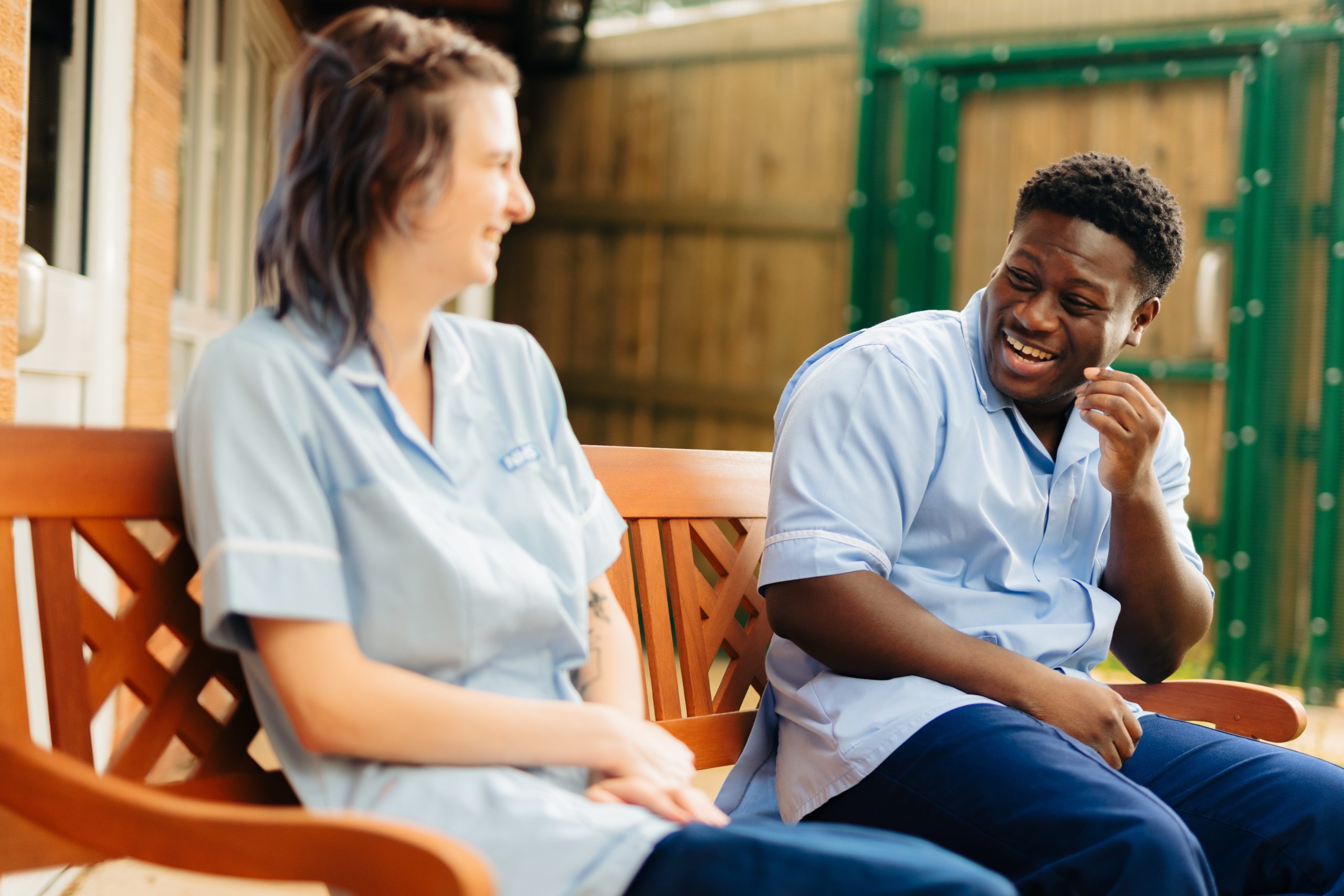 image of two colleagues sat on a bench laughing, both wearing health care uniforms